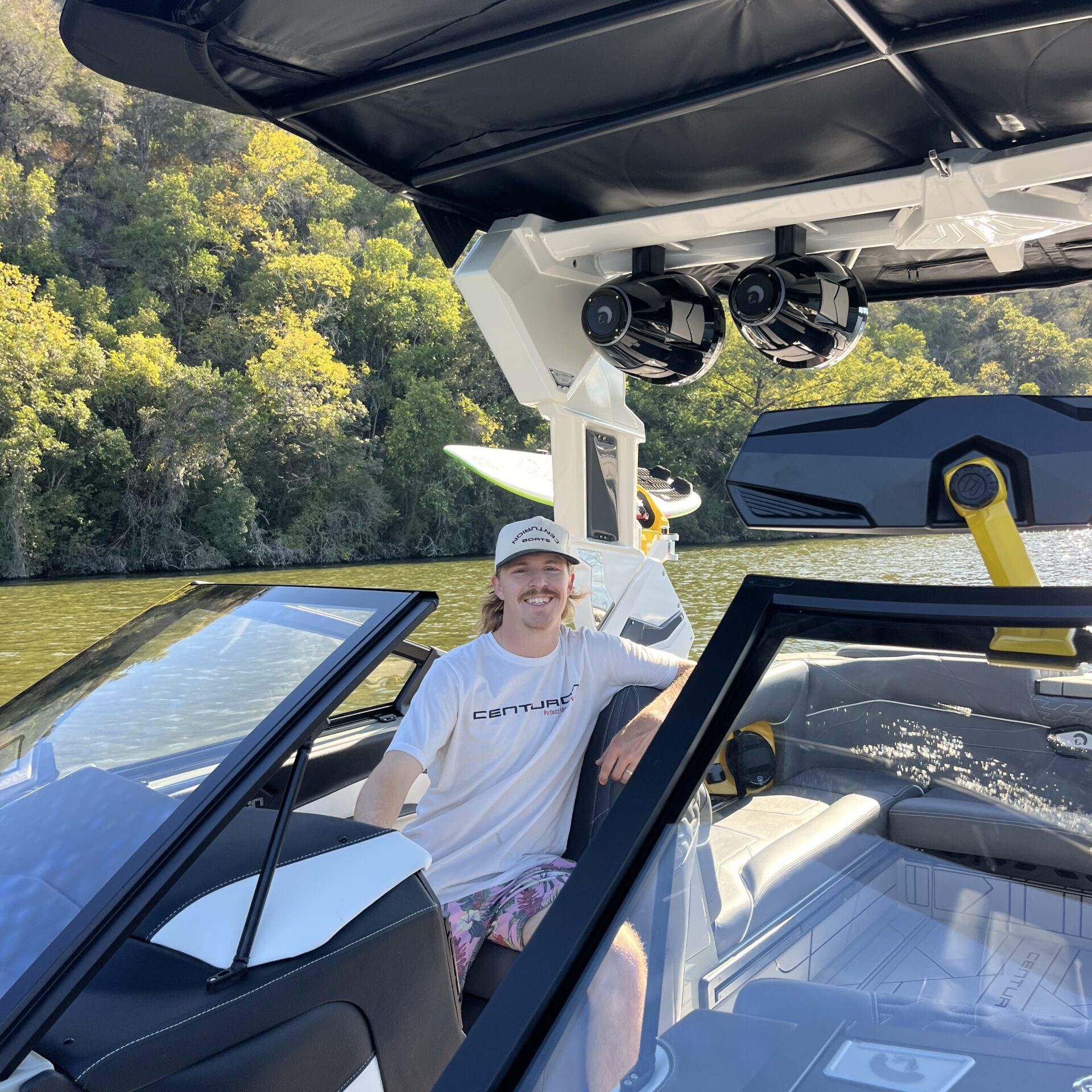 A person wearing a white shirt and cap sits at the helm of a boat on a lake, with trees and hills visible in the background.