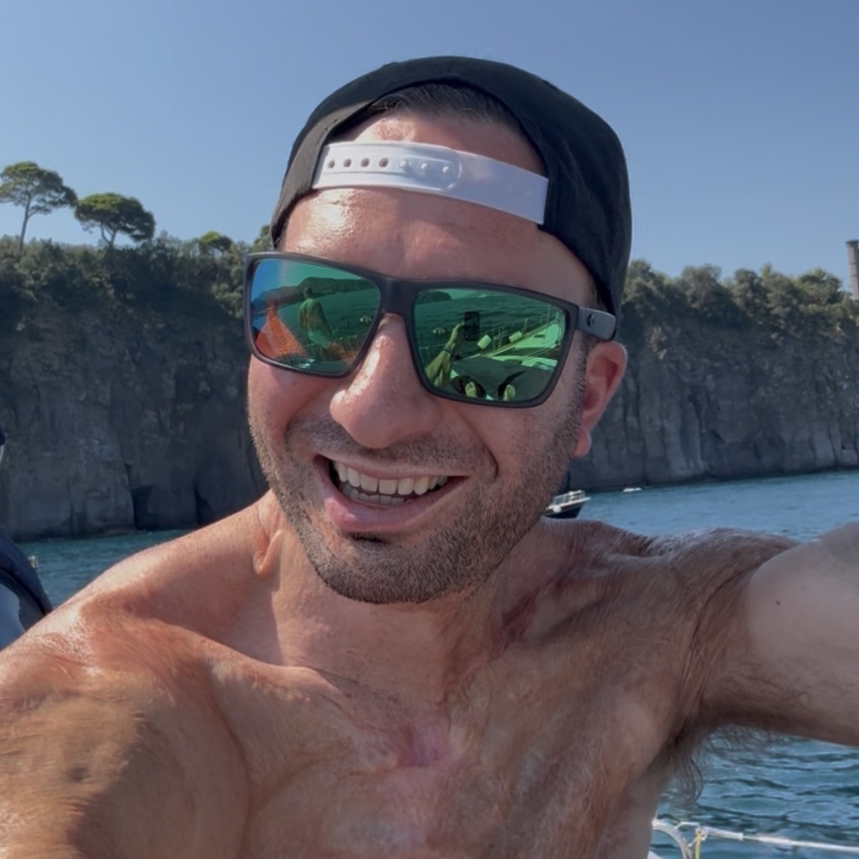 A man wearing sunglasses and a backward cap takes a selfie on a boat with rocky cliffs and water in the background on a sunny day.
