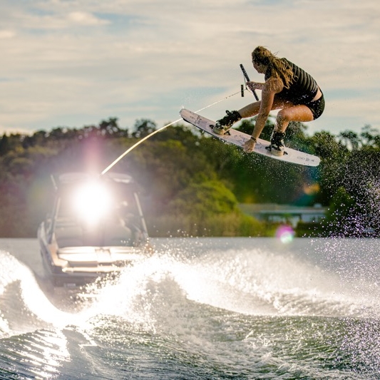 A person is wakeboarding, performing a jump and trick above the water, with a boat towing them in the background.