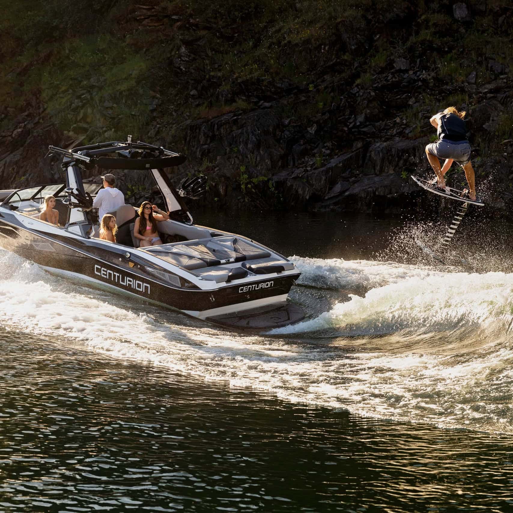 A person wakeboards and performs a jump behind a Centurion boat with several passengers on a sunlit lake near a rocky shoreline.