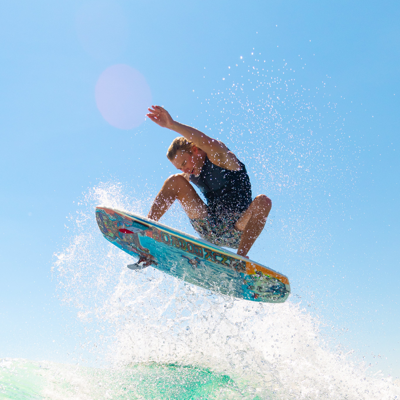 A person mid-air on a surfboard above a wave, with water splashing and a clear blue sky in the background.