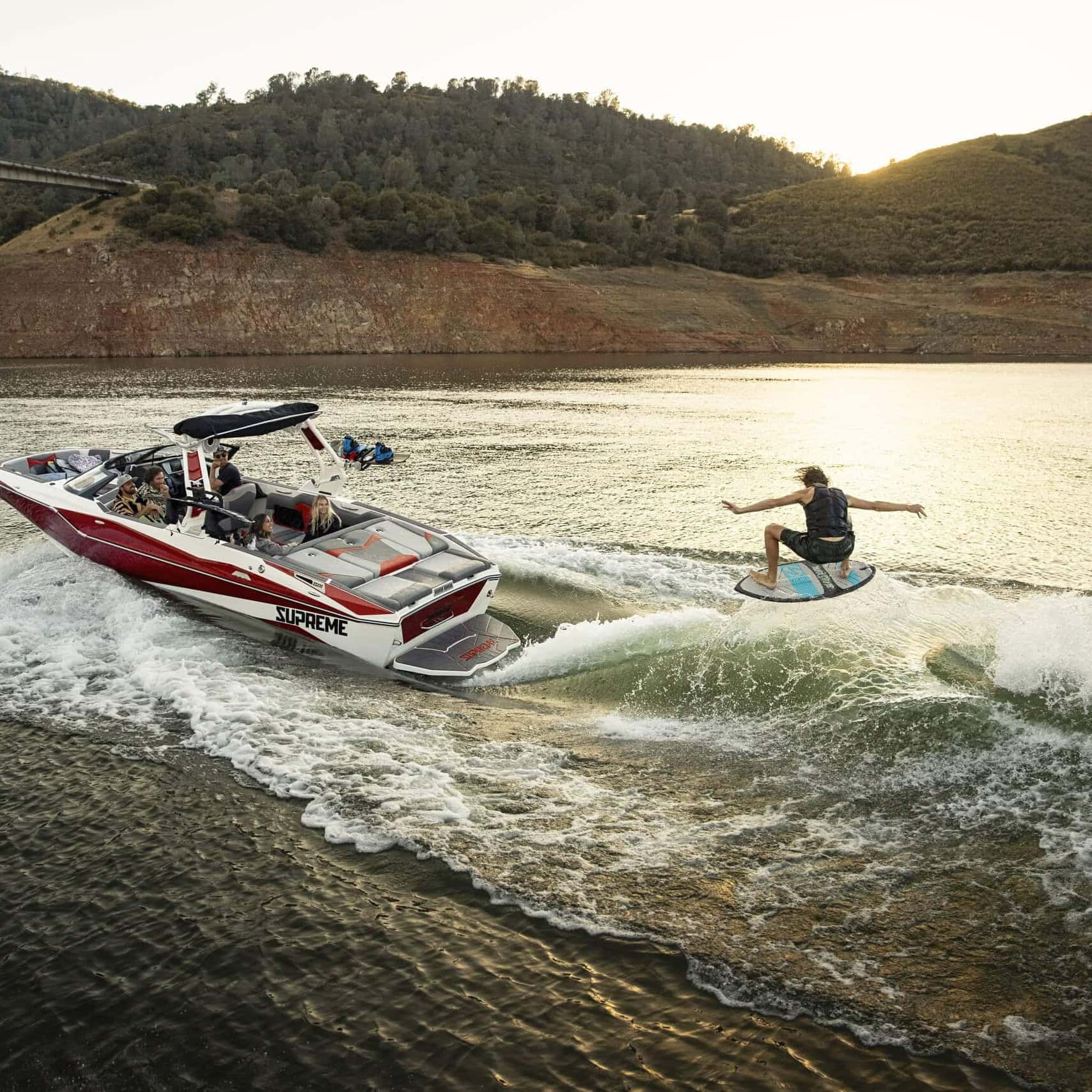 A person is wakesurfing behind a red and white speedboat on a lake, with hills and a bridge visible in the background at sunset.