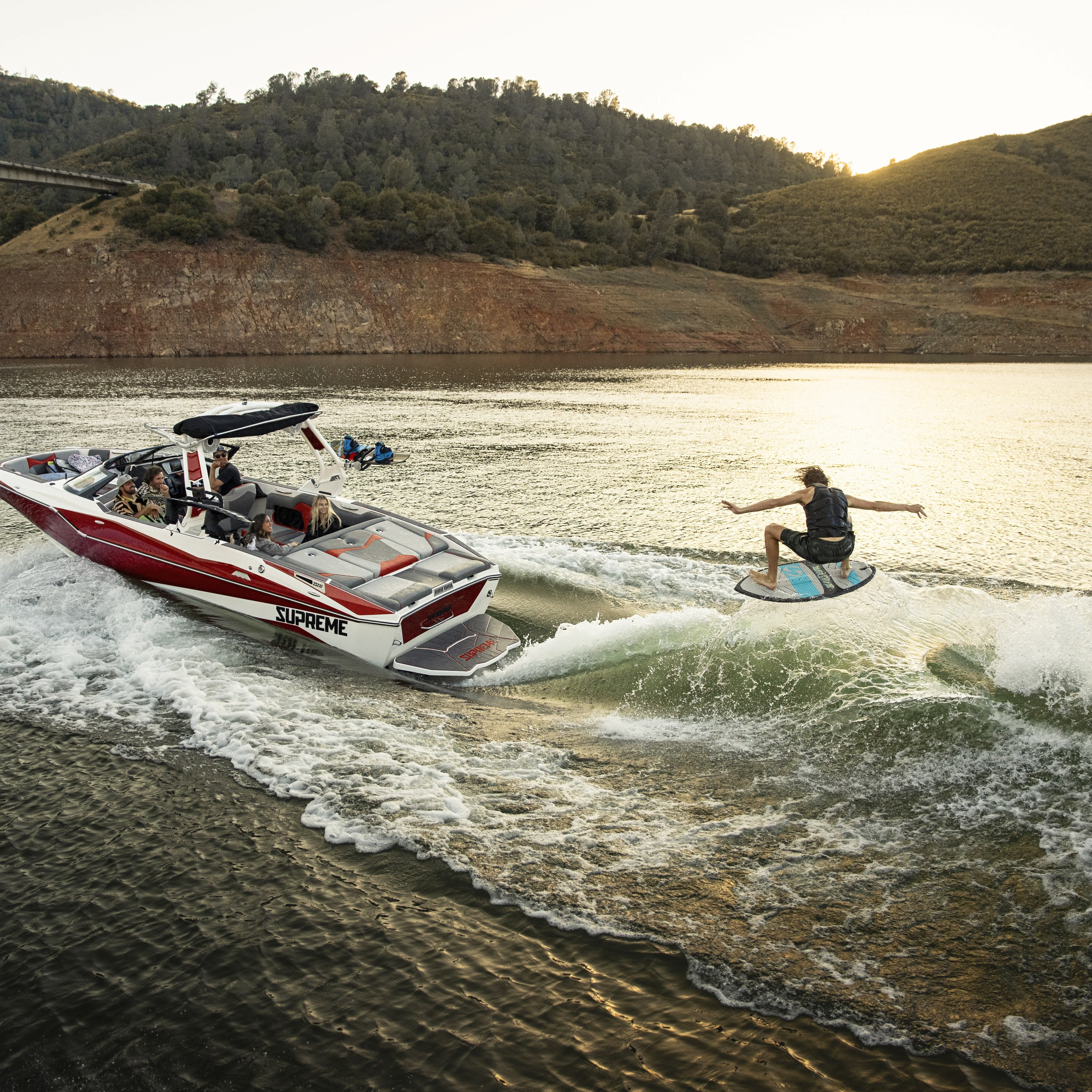 A person is wakesurfing behind a red and white speedboat on a lake, with hills and a bridge visible in the background at sunset.