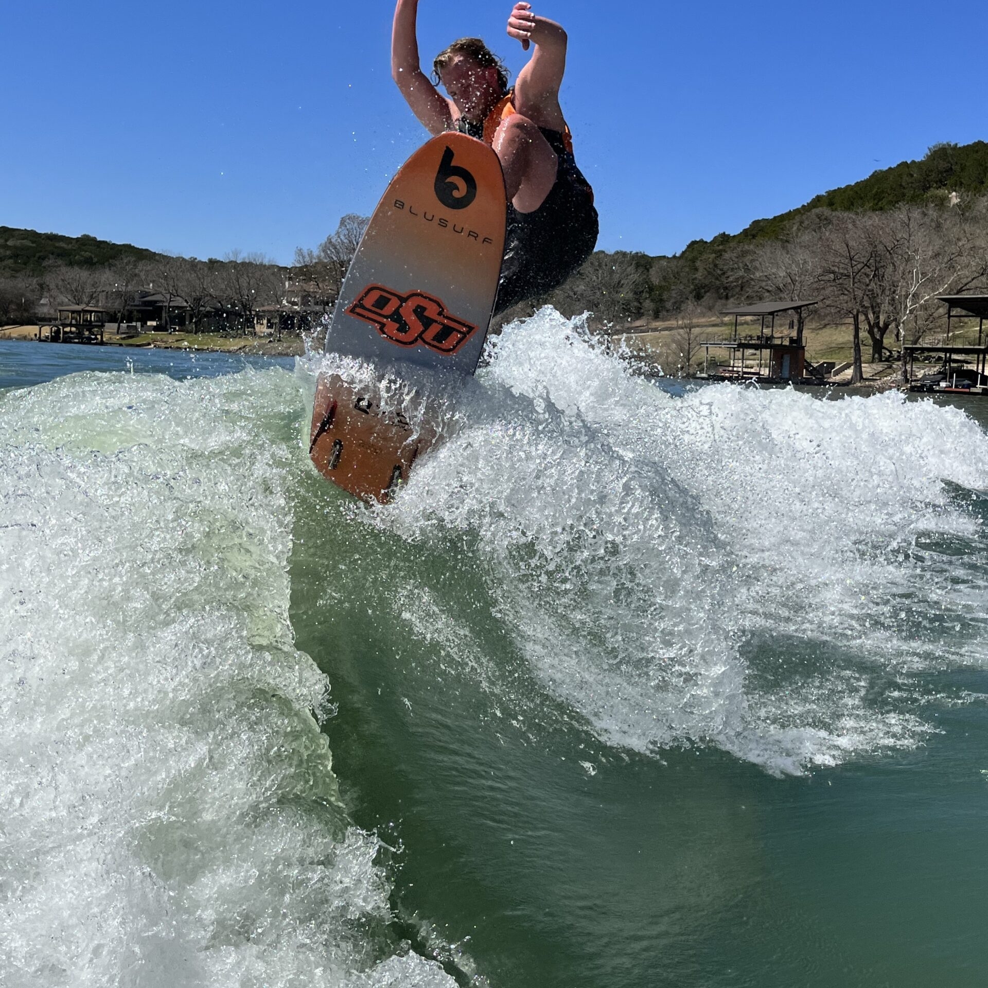A wakesurfer wearing a life vest performs a trick on a wave, with clear blue sky and lakeside houses visible in the background.