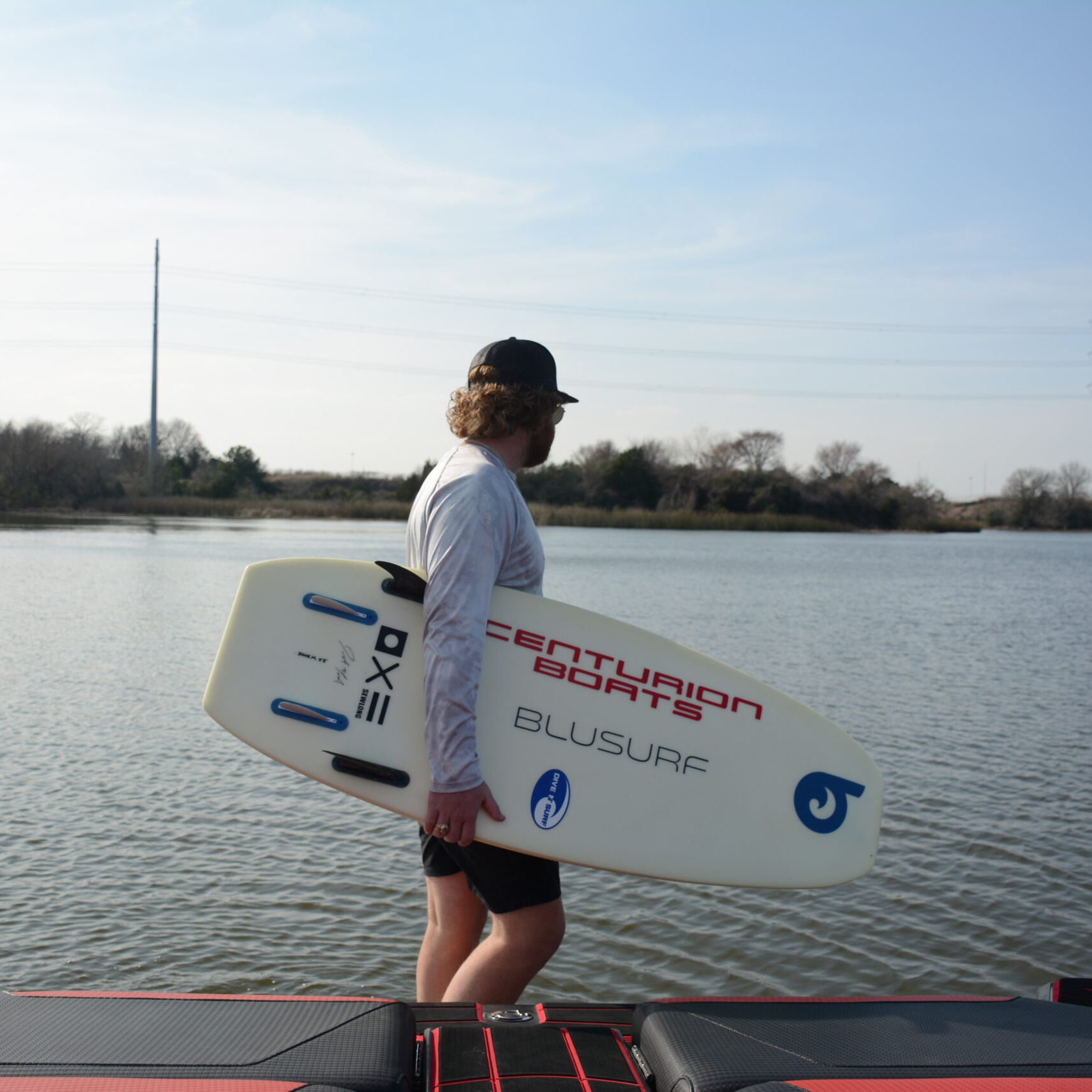 A person stands at the edge of a lake holding a white surfboard with branding, wearing a cap, sunglasses, and a long-sleeve shirt. Trees and power lines are visible in the background.