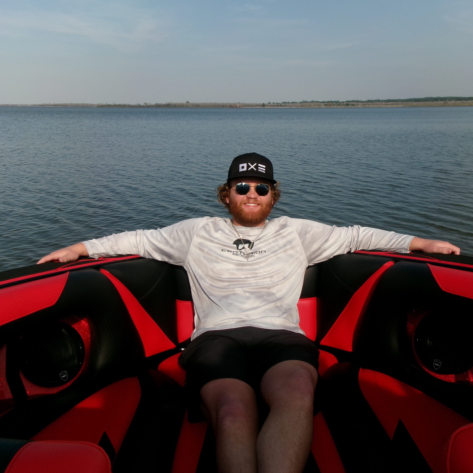 A man wearing sunglasses and a cap relaxes with arms outstretched on the red and black seats of a boat on a calm lake under a clear sky.