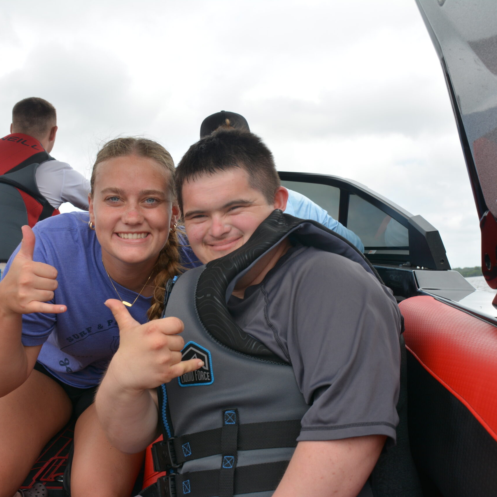Two young people wearing life jackets smile and make hand signs while sitting on a boat, with others in the background.