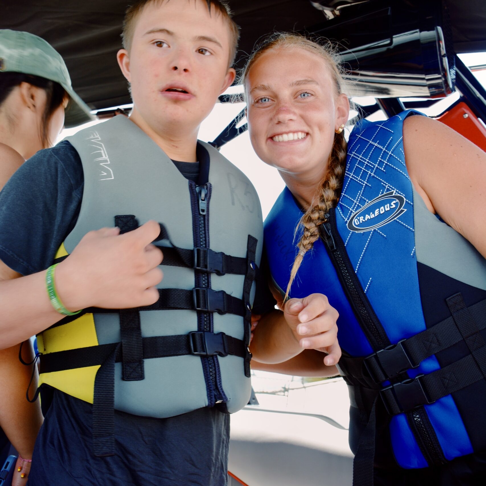 Two young people wearing life jackets stand on a boat; one looks ahead while the other smiles at the camera. Another person is partially visible in the background.