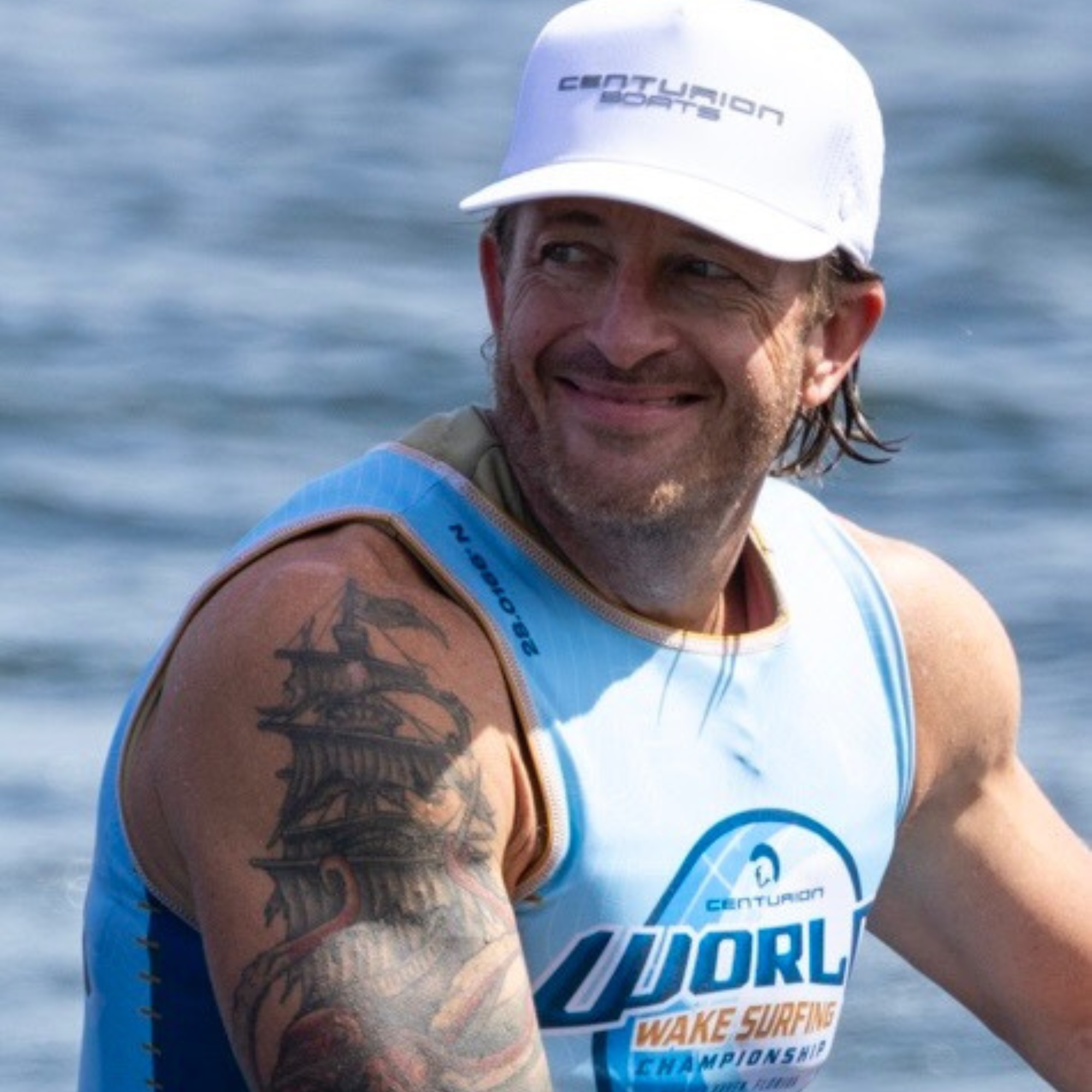 Chad Hudson, a wake surfer in a blue sleeveless shirt and white cap, sits by the water, smiling. A detailed ship tattoo stands out on his left arm.