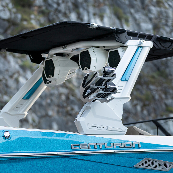 Close-up of a blue Centurion boat featuring a wakeboard tower with mounted speakers and board racks, with rocky cliffs in the background.