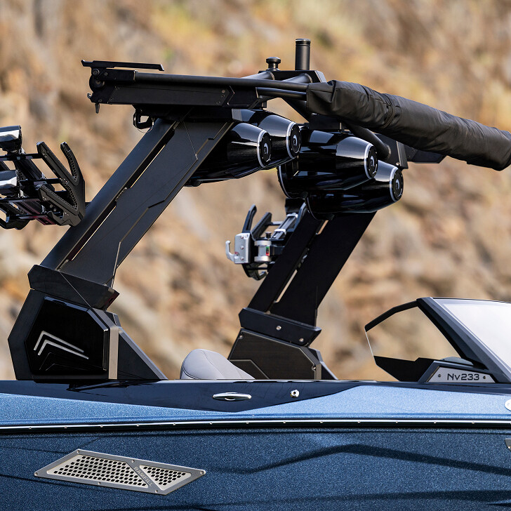 Close-up of a boat's tower with mounted speakers, wakeboard racks, and a folded bimini top; part of the blue hull and windshield are visible.