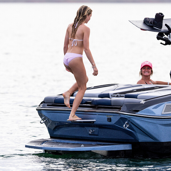 A woman in a pink bikini steps onto the back of a blue motorboat, while another woman in a pink cap sits and smiles in the background.