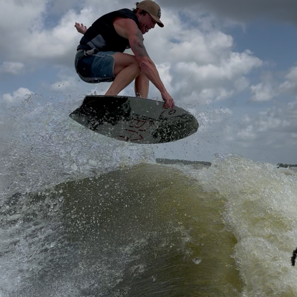 Chad Hudson, a skilled wake surfer wearing a life vest and cap, performs an airborne trick on his wakesurf board above a wave, with splashing water and a cloudy sky in the background.
