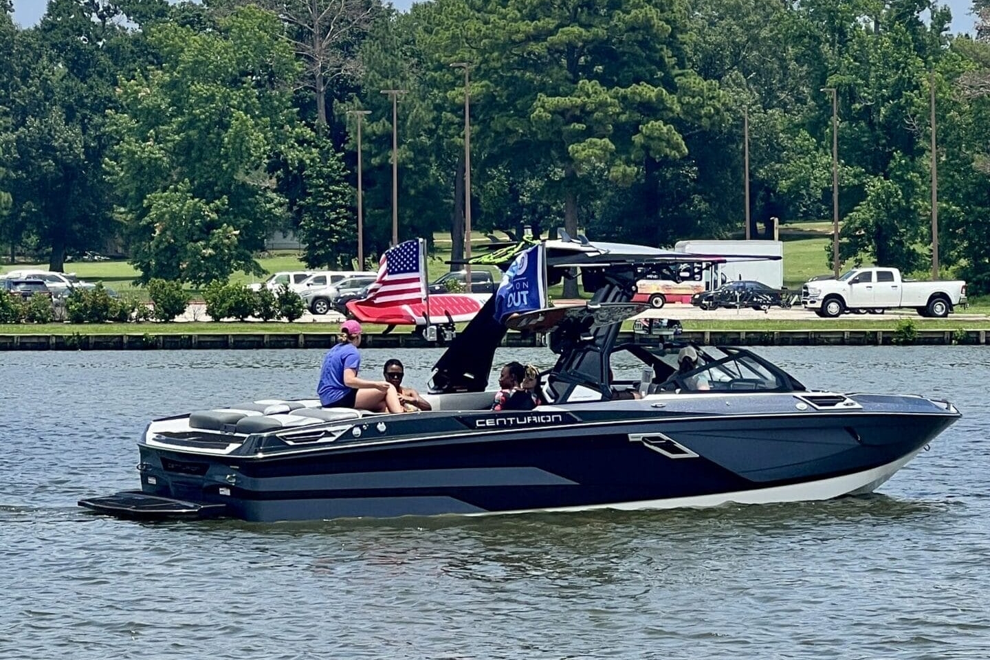 A Centurion Boats Salute to Service event honors heroes at the Come and Take it Wake Surf Classic in Lake Conroe, where a motorboat with several people on board navigates a calm lake. An American flag is visible on the boat, and trees and parked vehicles line the shoreline in the background.