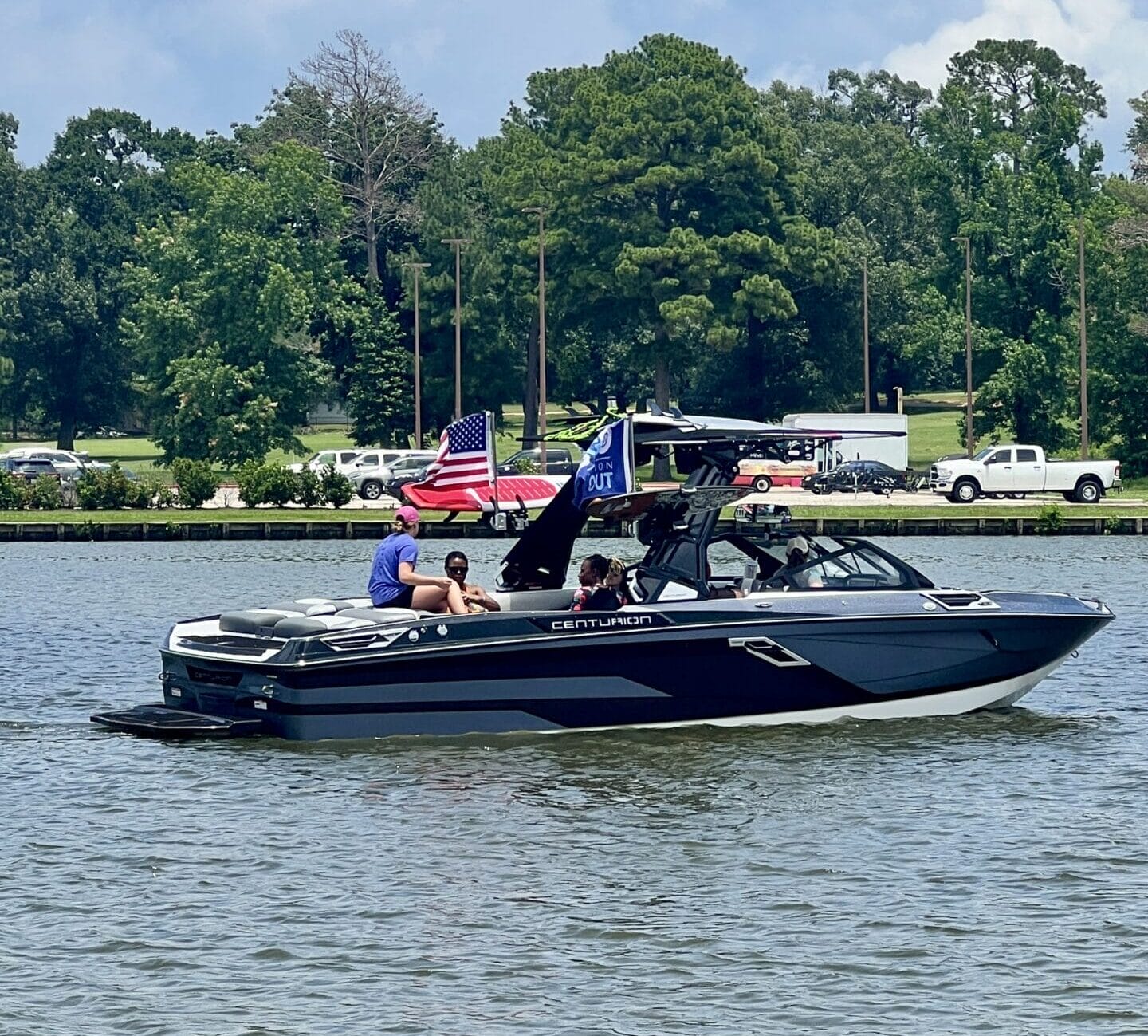 A Centurion Boats Salute to Service event honors heroes at the Come and Take it Wake Surf Classic in Lake Conroe, where a motorboat with several people on board navigates a calm lake. An American flag is visible on the boat, and trees and parked vehicles line the shoreline in the background.