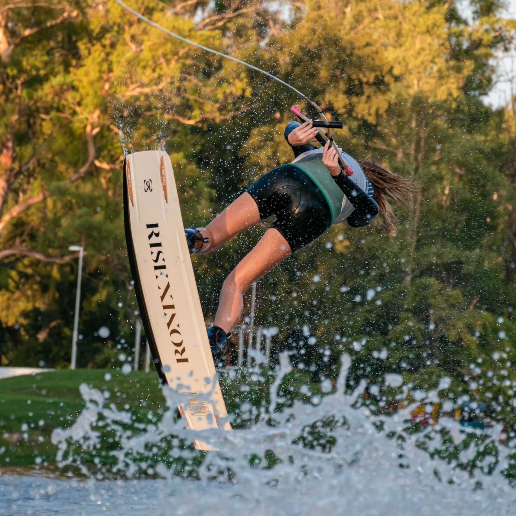 Kitt Smith is wakeboarding, catching air above the water while holding onto a tow rope, with trees and grass visible in the background.