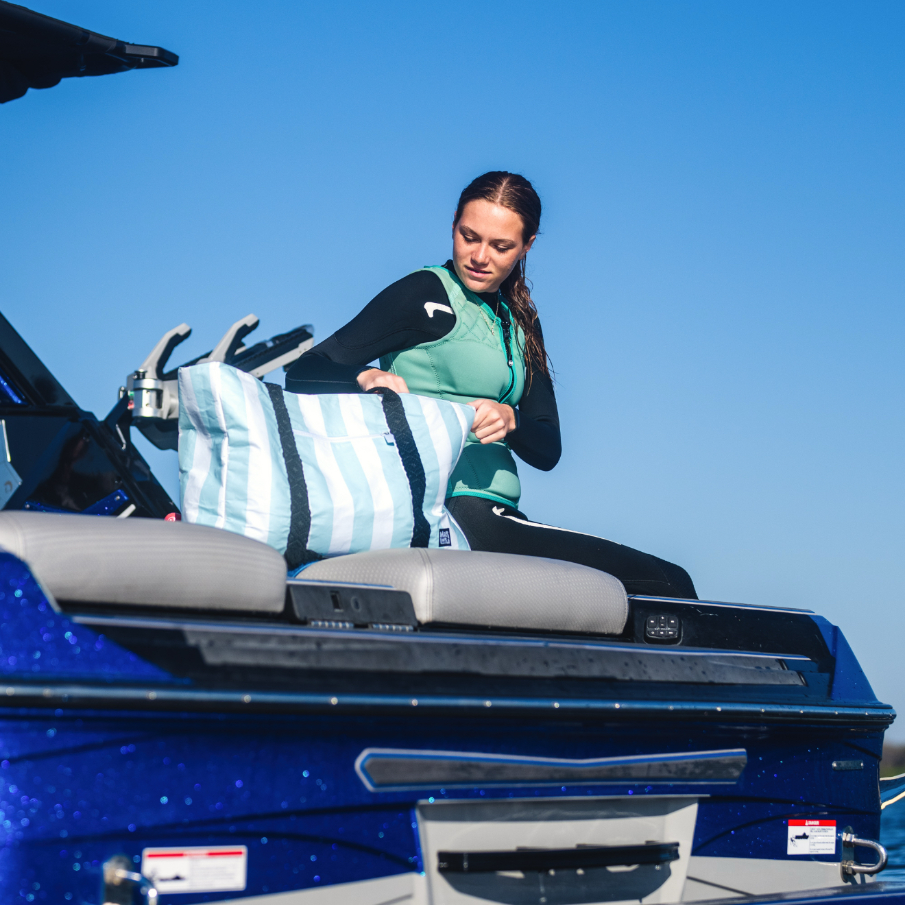 Kitt Smith, in a wetsuit, sits on a blue boat, placing a striped bag on the seat, with the clear blue sky in the background.
