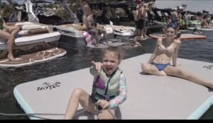 A young girl wearing a life jacket points toward the camera while sitting on a floating mat. Other people are relaxing on boats and in the water around her.