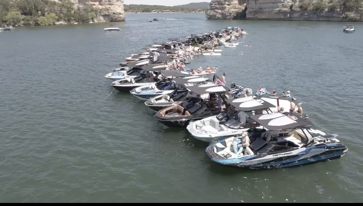 A large group of boats is lined up on a lake, forming a semi-circle. The boats are filled with people enjoying the water and sunny weather, with cliffs and trees visible in the background.