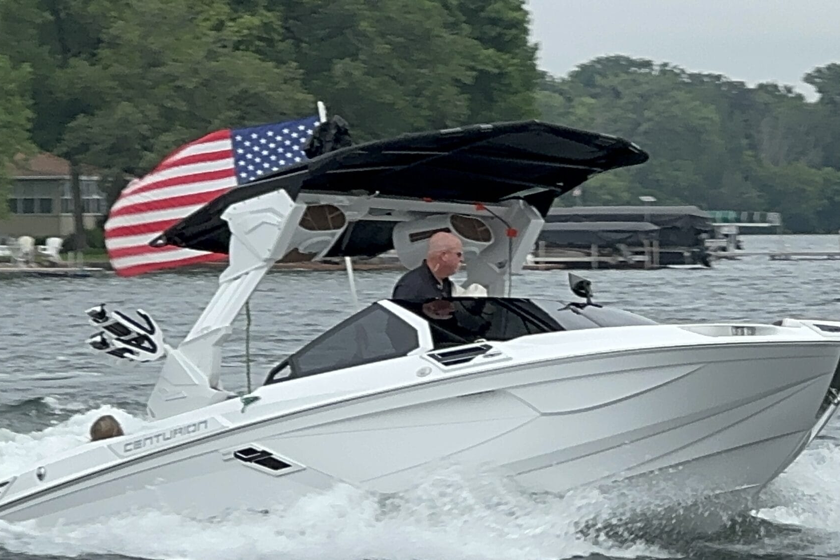A person is driving a white Centurion boat on a lake. The boat has an American flag at the back, and there are trees and houses visible in the background.