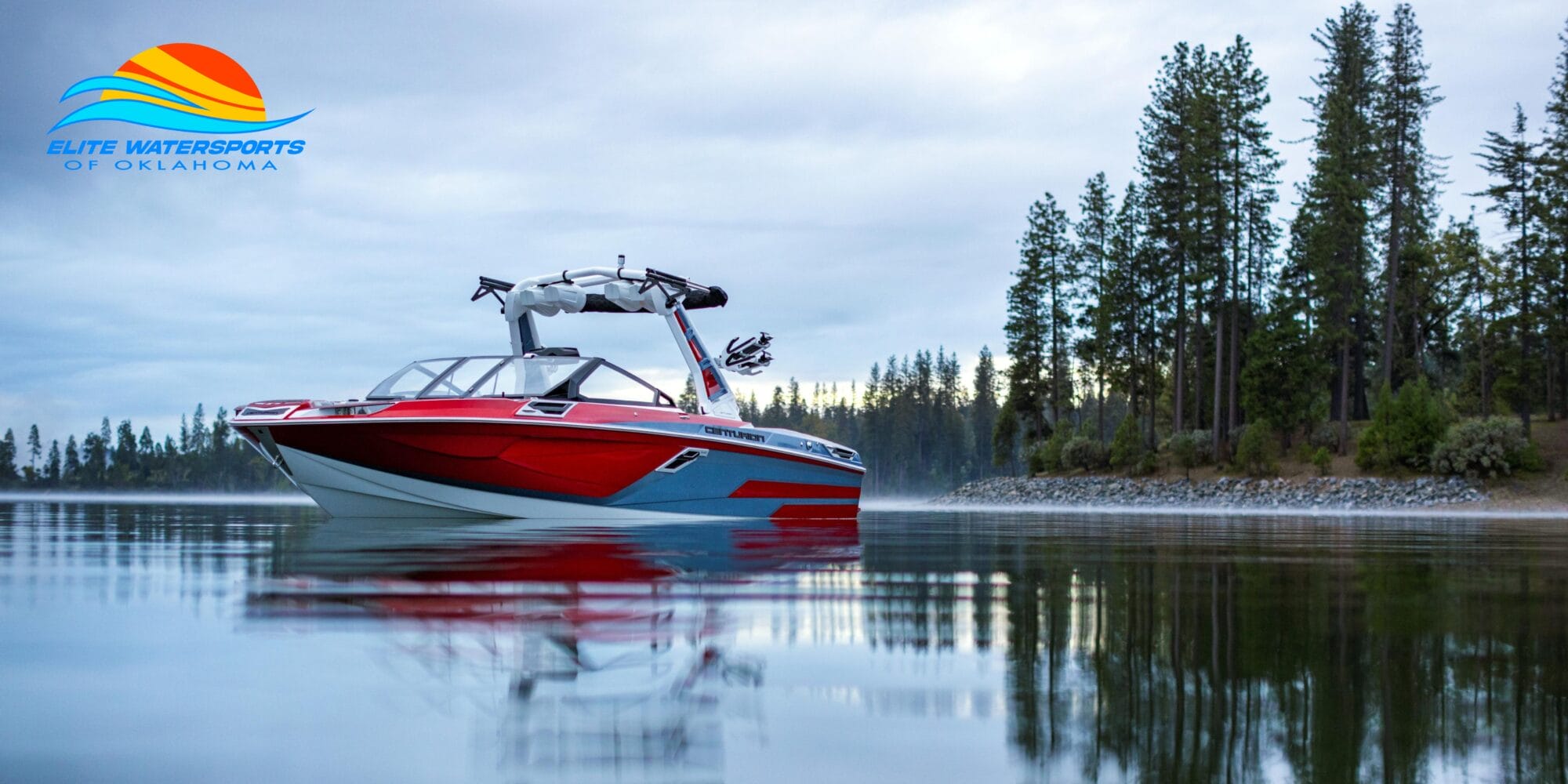 A red and white wakeboard boat is on a calm lake surrounded by trees, with a logo of "Elite Watersports of Oklahoma" in the top-left corner.