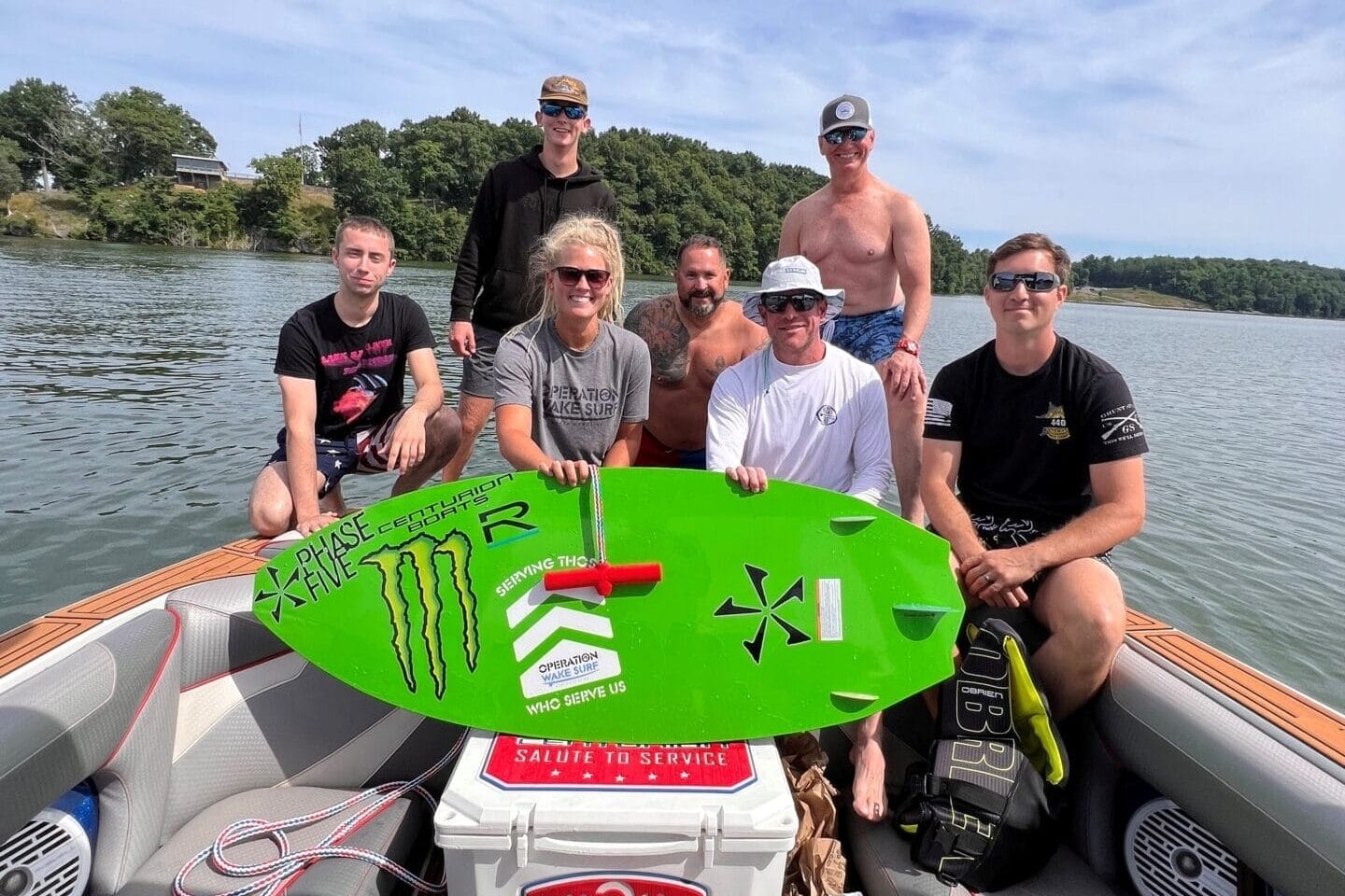 A group of seven people on a boat, one holding a green surfboard. They are on a body of water with trees in the background, under a partly cloudy sky.