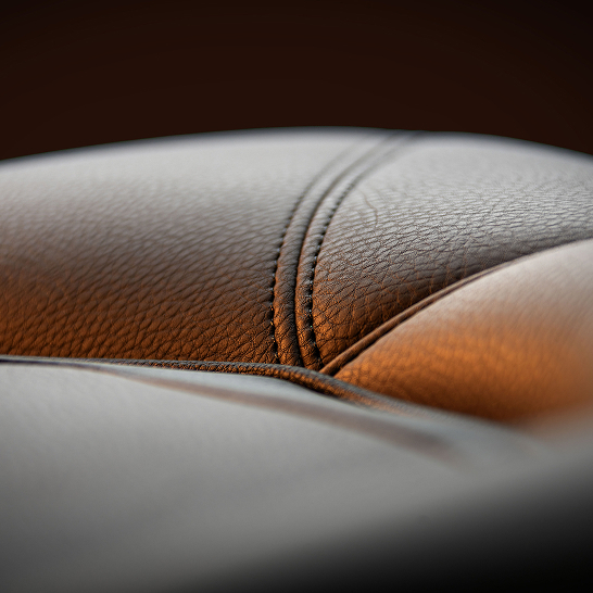 Close-up of stitched, textured brown leather upholstery from the Ri Series, with soft lighting highlighting the curved seams. Features Personal detailing for a refined finish.