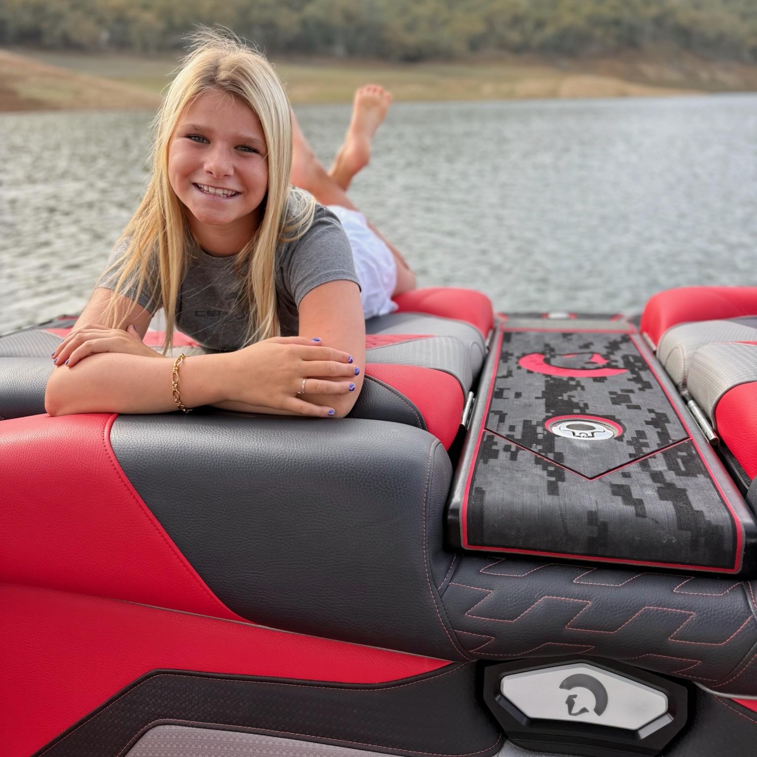 A young girl with long blonde hair lies on her stomach on the back seat of a red and gray Centurion Wake Surfer boat, smiling at the camera, with a lake and wooded hills in the background. Brynlee Hall enjoys a perfect day outdoors.