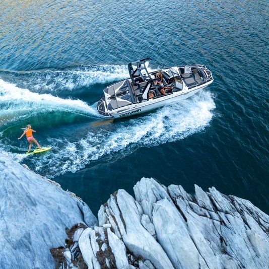 A person, possibly Centurion Wake Surfer Brynlee Hall, wakeboarding behind a motorboat on a lake, with rocky cliffs visible in the foreground.