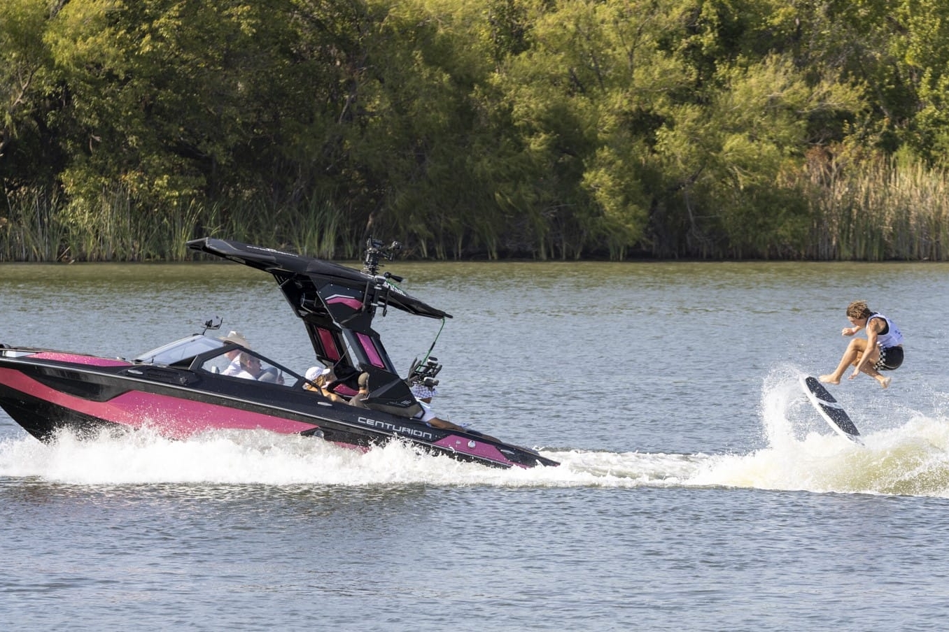 A person is wakeboarding behind a speedboat on a lake with trees in the background.