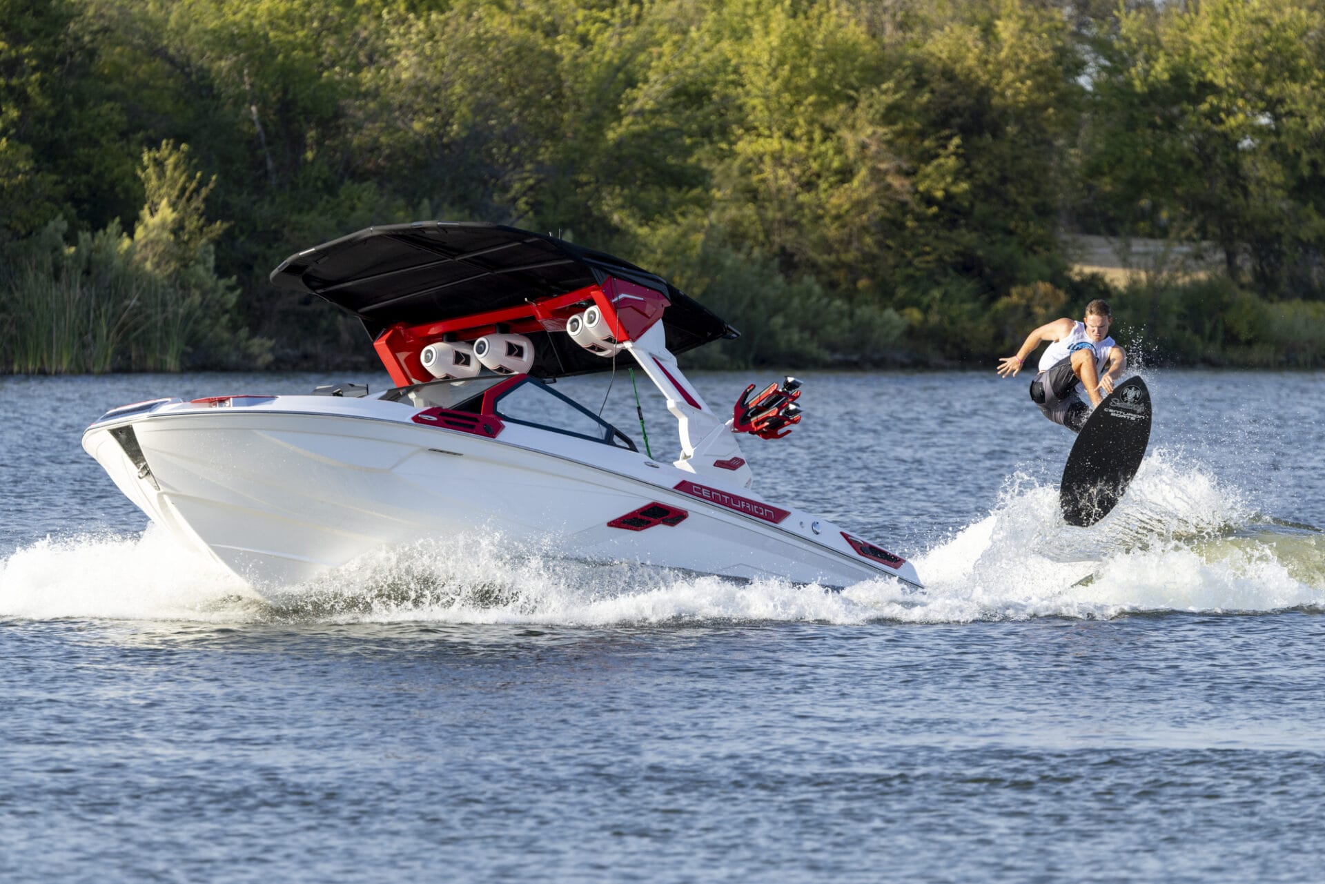 A person wakeboarding performs a trick in the air beside a moving motorboat on a lake with trees in the background.