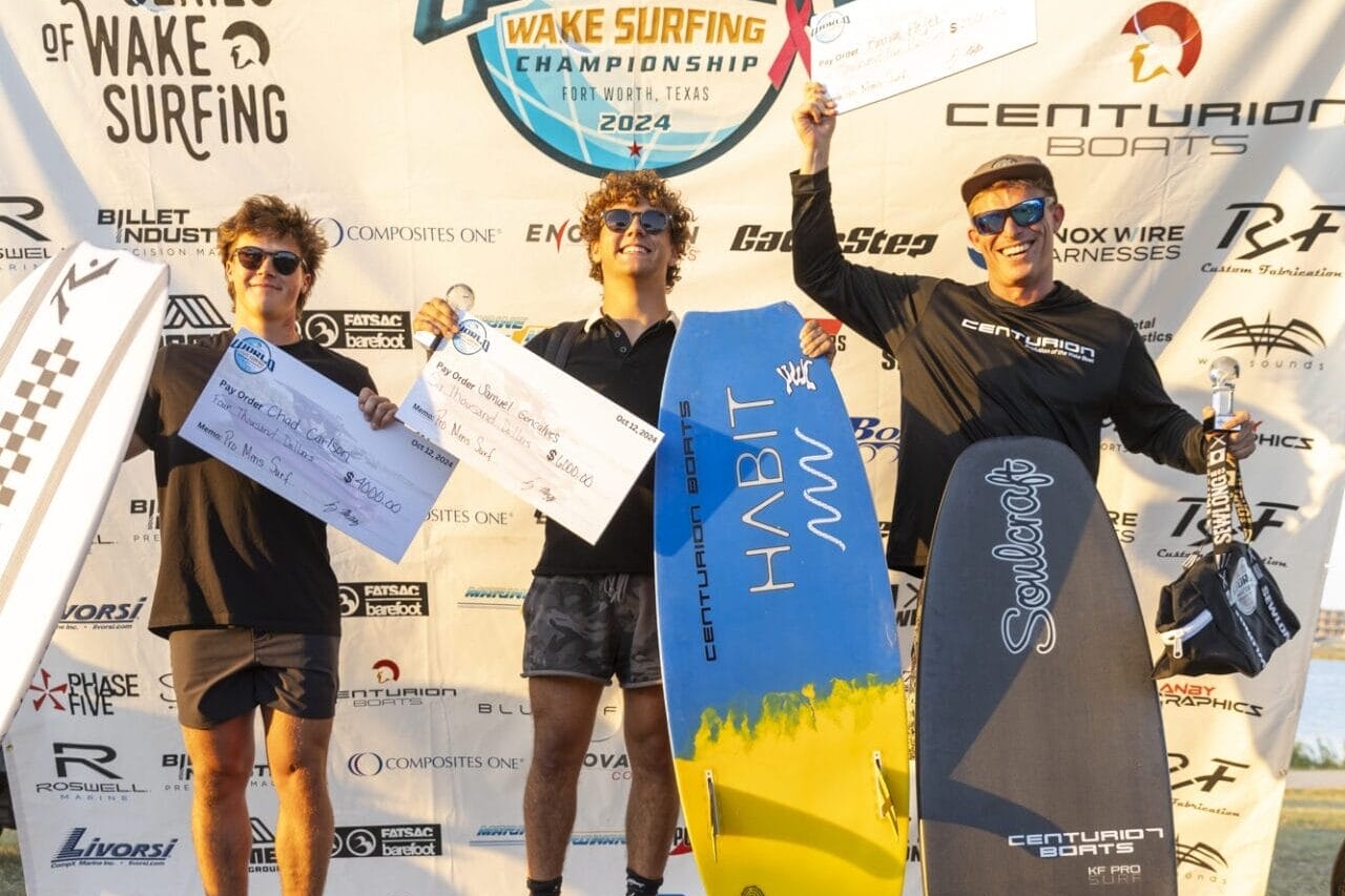 Three people stand on a podium holding surfboards and prize checks at a wake surfing championship event.