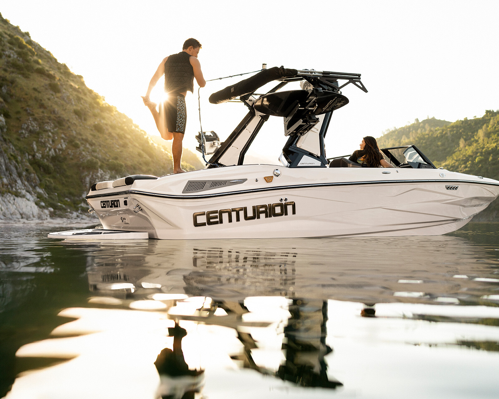 A man stands on the rear platform of a Centurion Nv213 Surf Boat, while a woman sits inside. The boat floats on calm water near rocky hills at sunset.