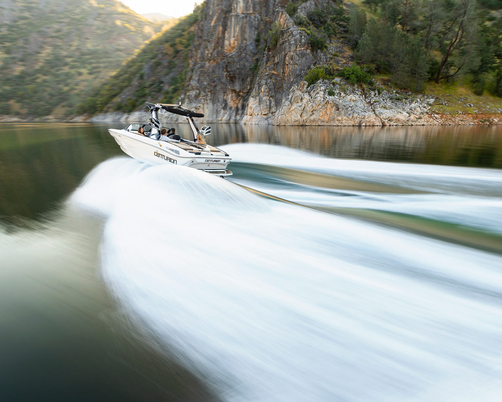 A Centurion Nv213 speeds across a calm lake, creating wide, blurred wake trails behind it with rocky hills and trees in the background.
