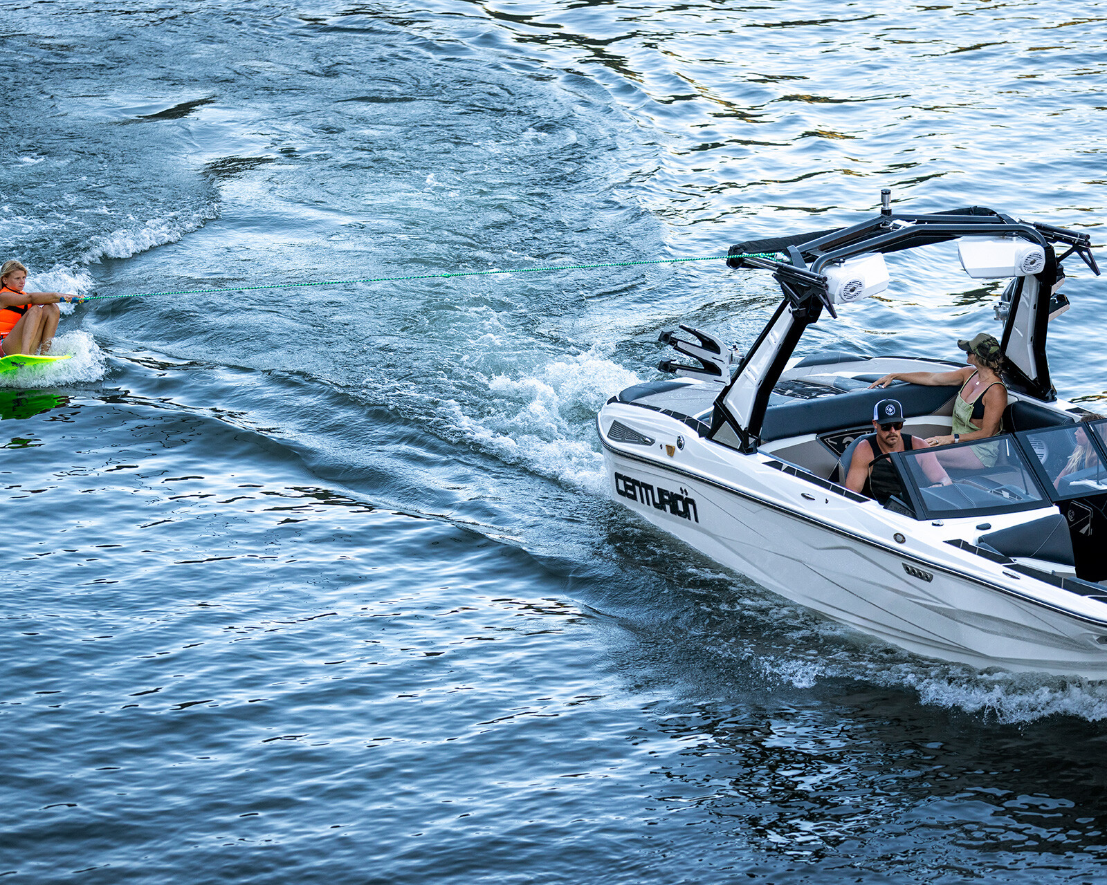 A person on a wakeboard is being towed by a white Centurion Nv213 surf boat across rippling water, with two people visible in the boat.