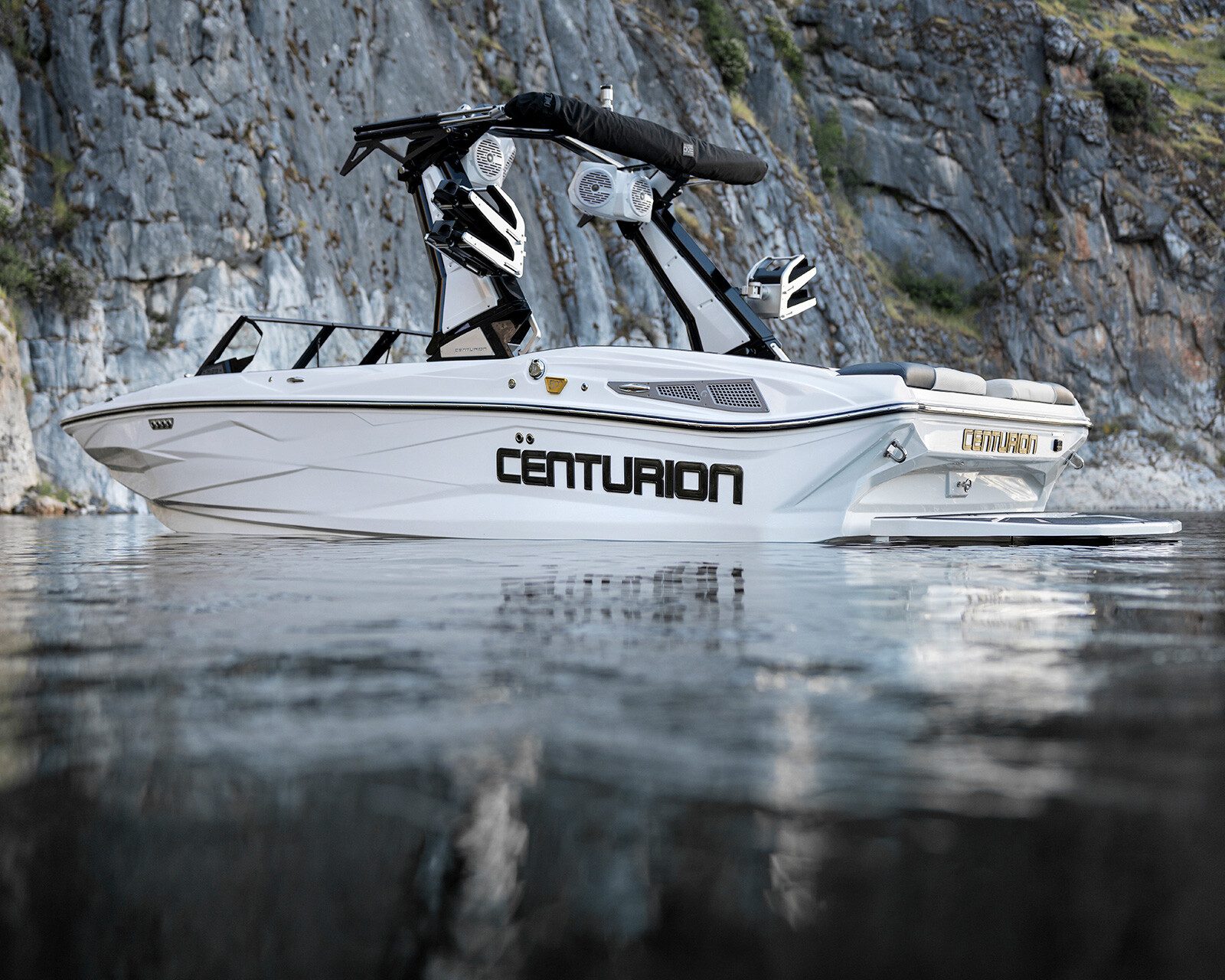 A white Centurion Nv213 Surf Boat floats on calm water near a rocky cliff, with wakeboard racks and speakers mounted above the seating area.