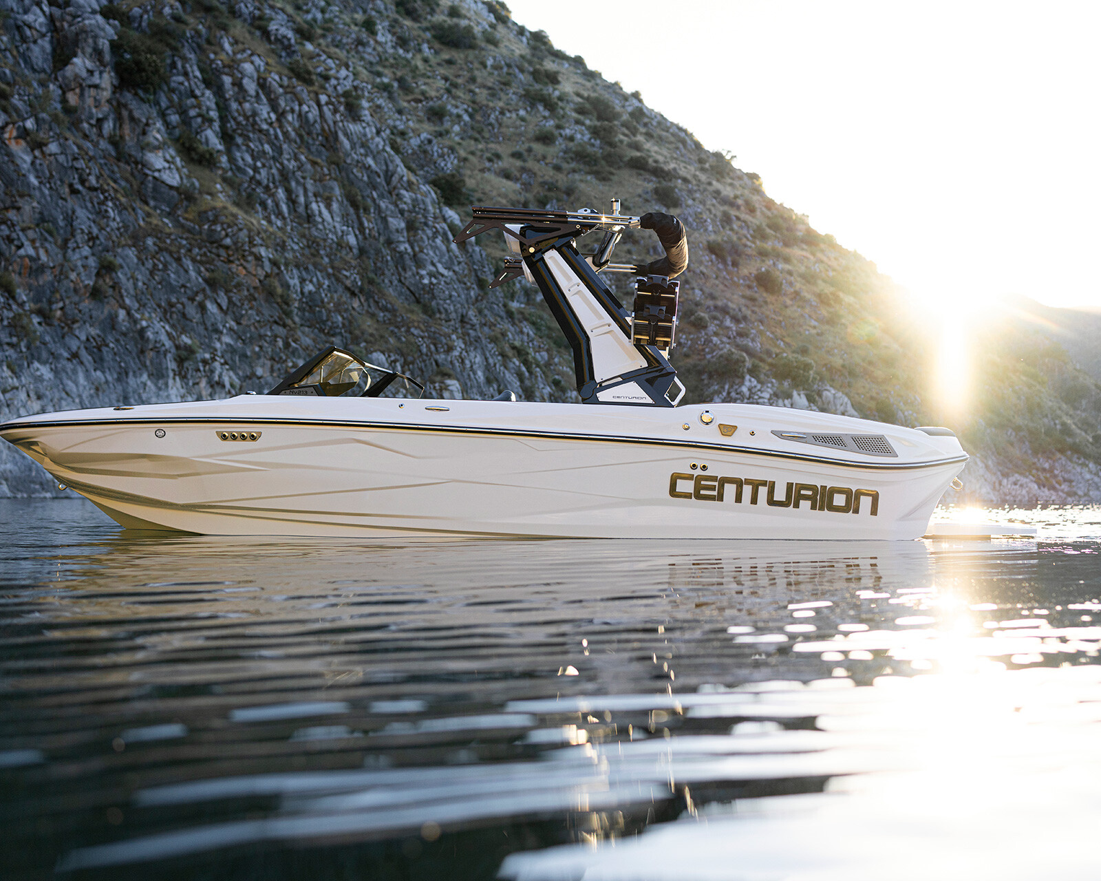 A white Centurion Nv213 speedboat floats on calm water near rocky hills at sunset.