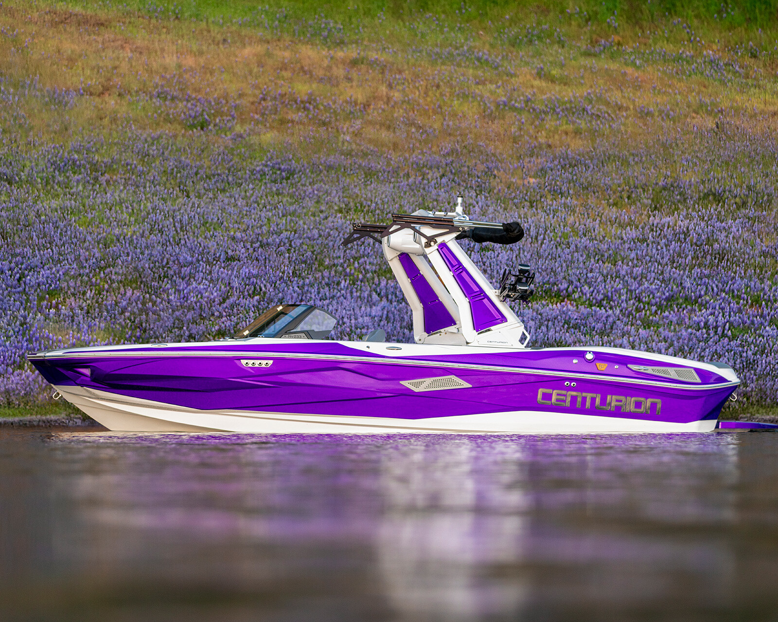 A purple and white Centurion Nv233 wake boat floats on calm water, with a grassy, flower-covered hillside in the background.