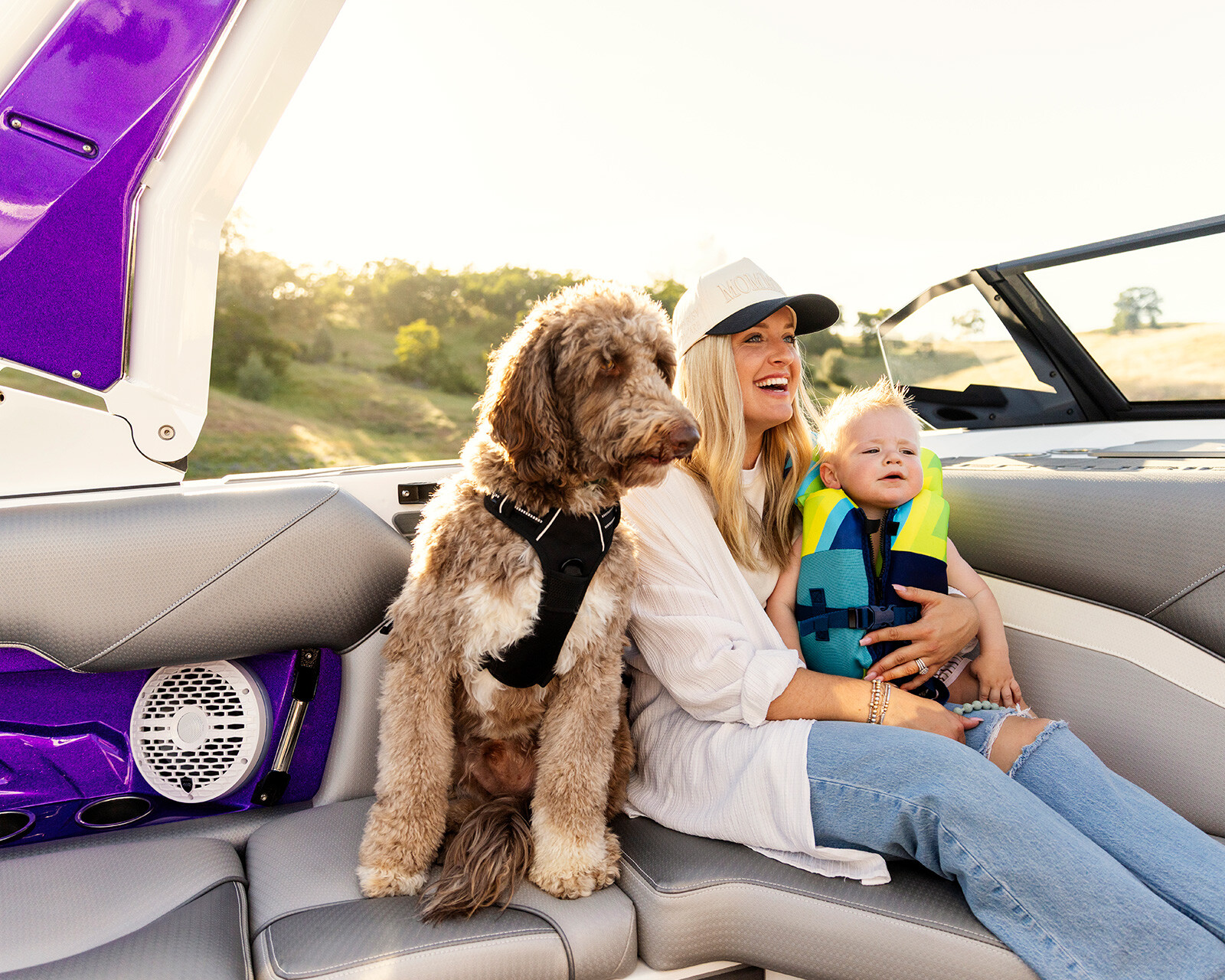 A woman, a baby wearing a life jacket, and a dog sit together on the back seat of a Centurion Nv233, with sunlight and trees in the background.