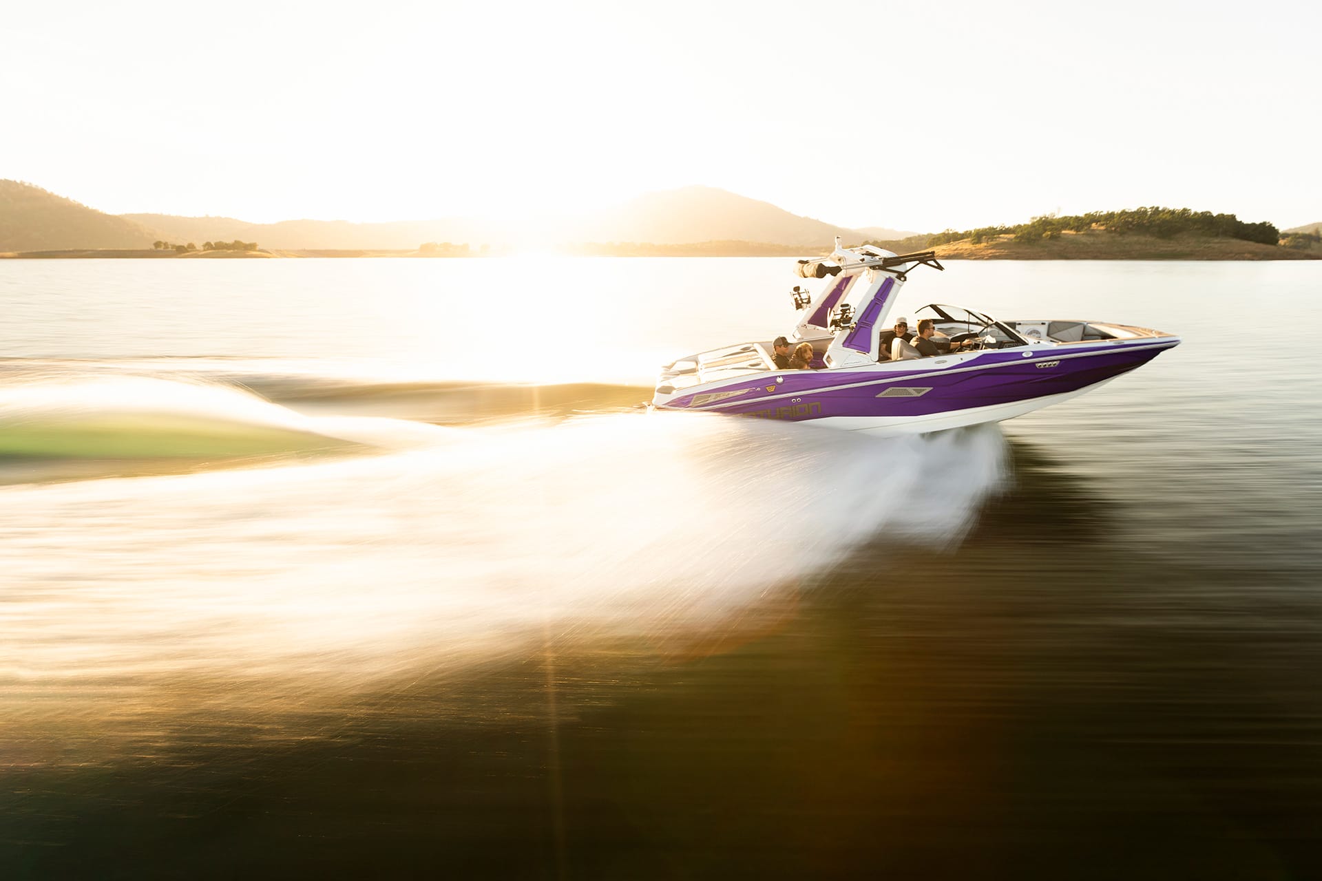 A purple Nv233 Wake Boat moves quickly across a calm lake, leaving a trail of white water, with mountains and the sun in the background.