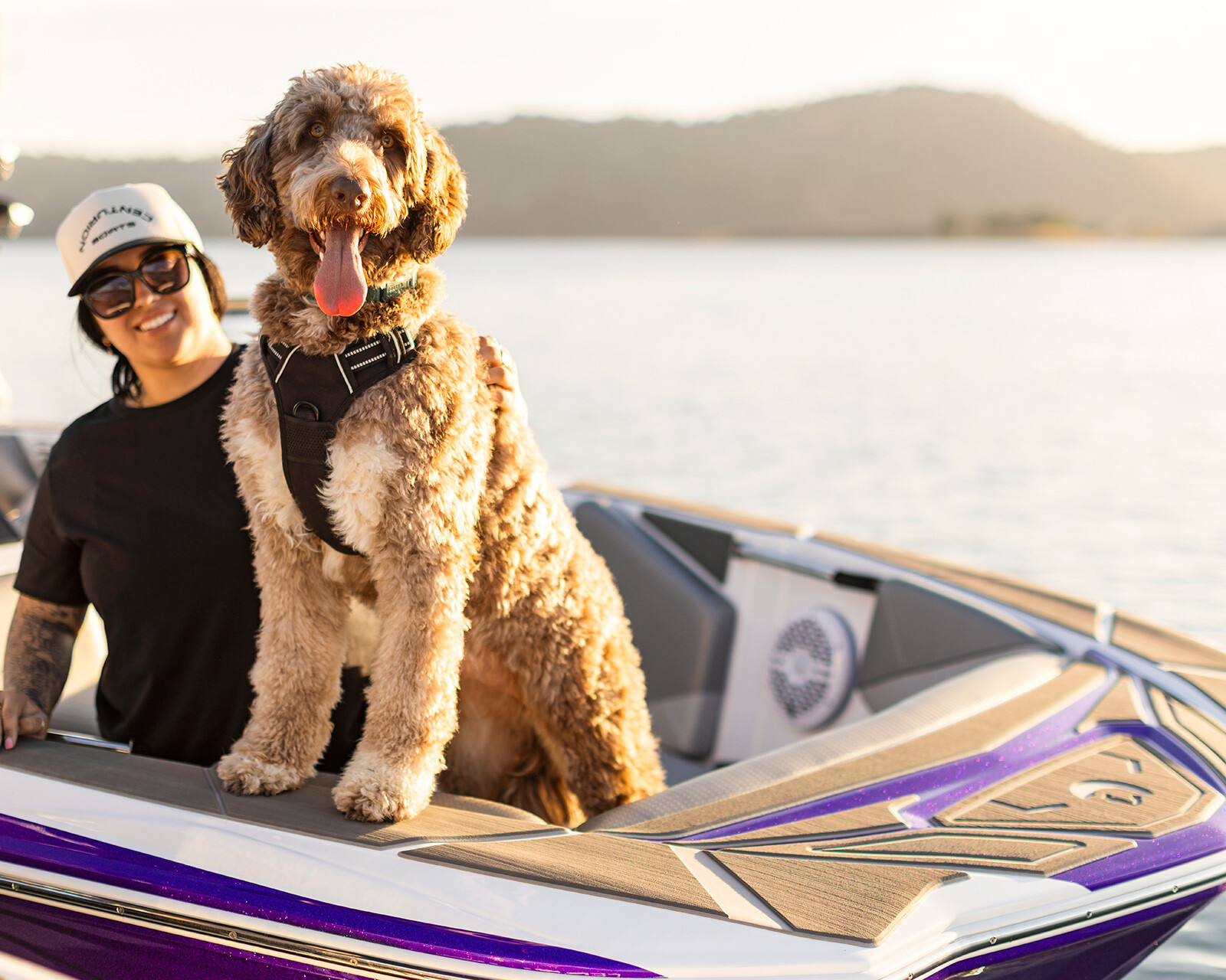 A woman wearing sunglasses and a cap sits on a Centurion Nv233 boat next to a large brown and tan dog with a harness. Water and hills are visible in the background.