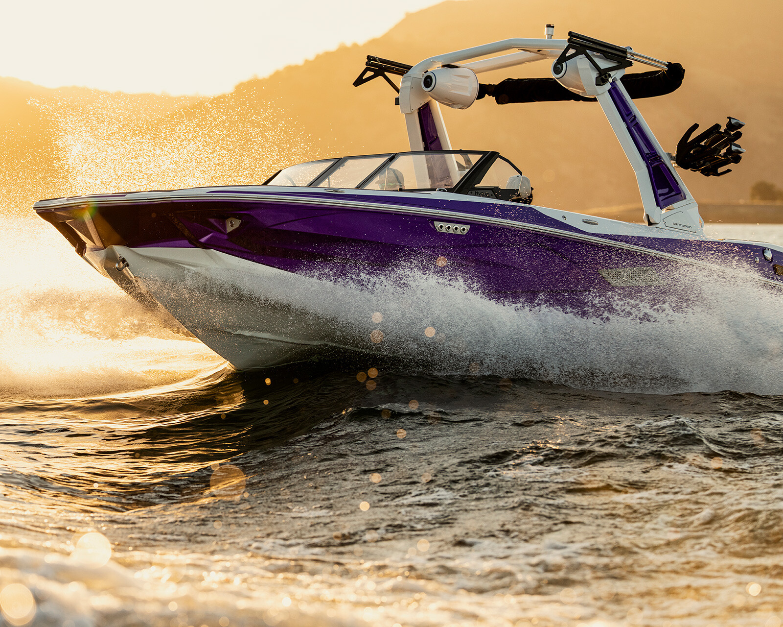 A purple and white Centurion Nv233 speedboat moves quickly across the water, creating splashes, with hills and the sun visible in the background.