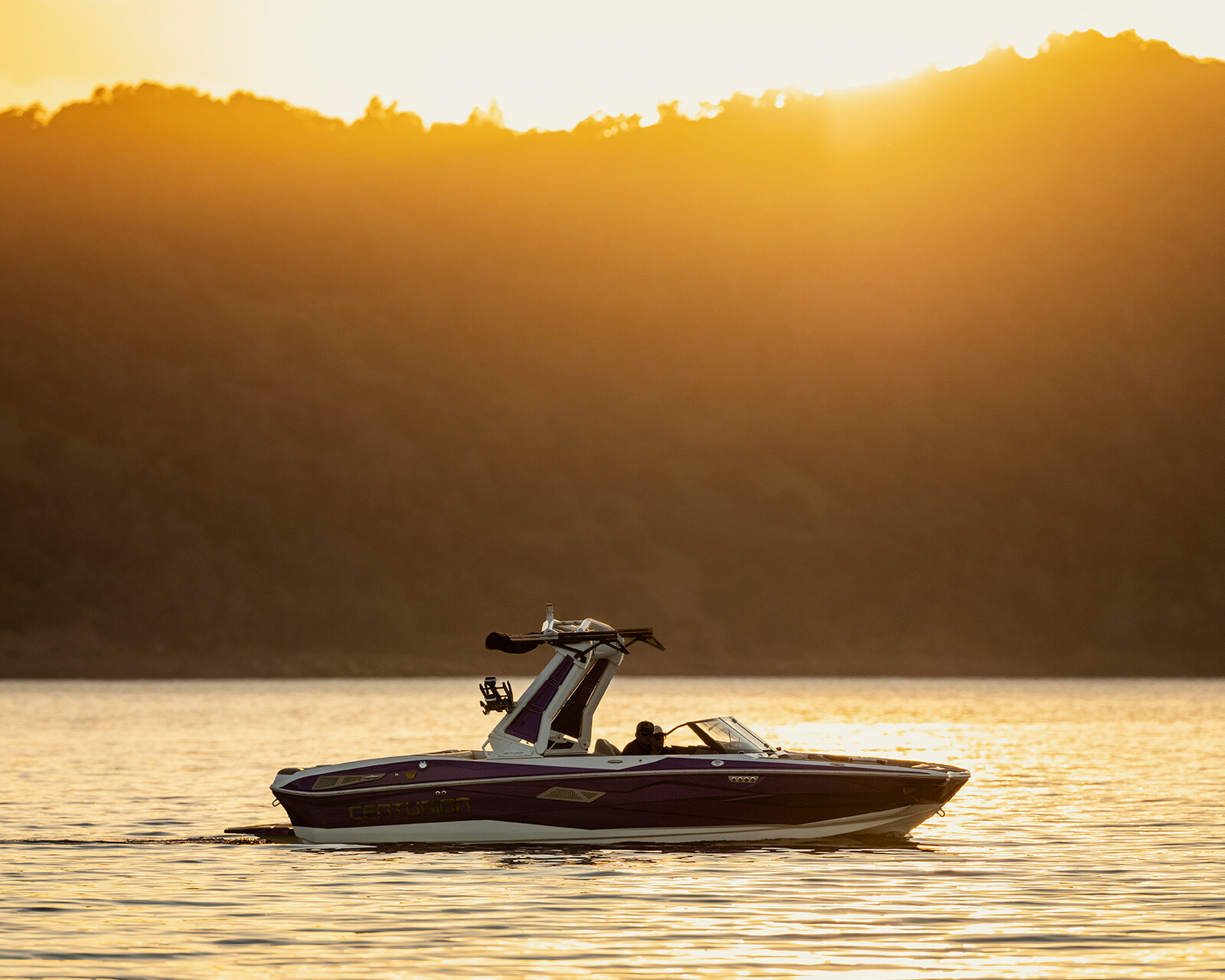 A Centurion Nv233 glides across a calm lake at sunset, the sun setting behind a silhouette of tree-covered hills.