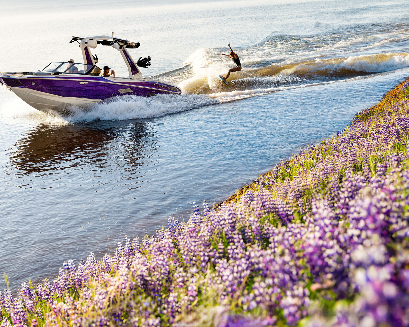 A Nv233 Wake Boat pulls a person wakeboarding on a calm river bordered by a field of blooming purple wildflowers.