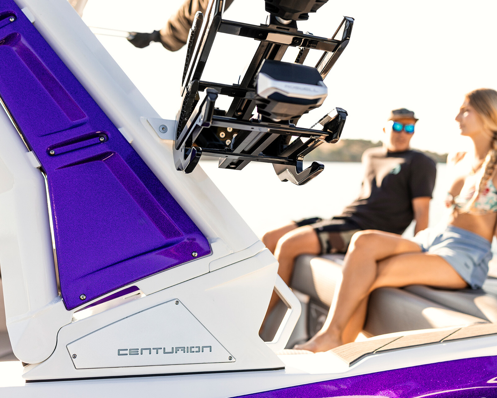 Close-up of a purple and white Centurion Nv233 Wake Boat with two people sitting and talking in the background on the water.