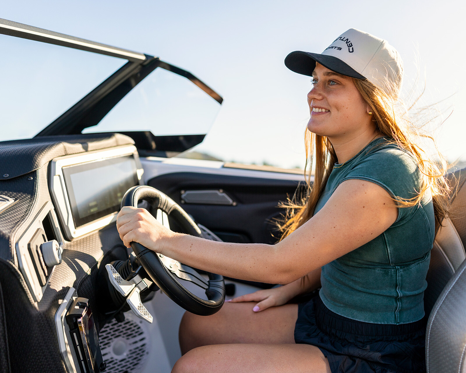 A woman wearing a cap and T-shirt is smiling while driving a Centurion Nv233 surf boat on a sunny day.