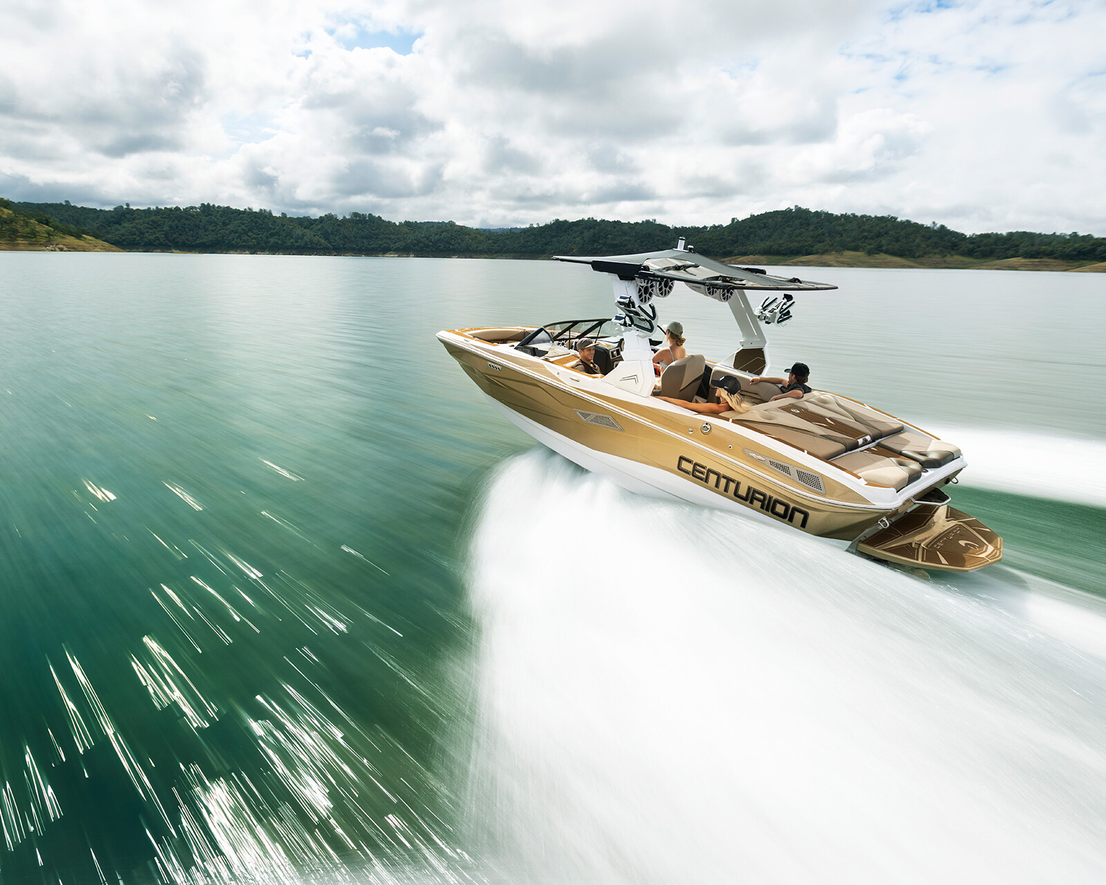 A gold Centurion Nv213 wake boat with several people onboard moves quickly across a calm lake, leaving white wakes behind, with forested hills in the background.