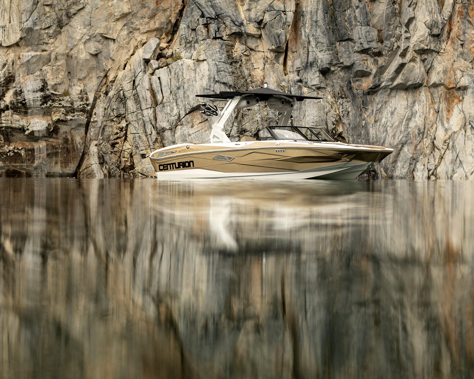 A white and gold Centurion Nv213 floats on calm water in front of a rocky cliff, with the boat and cliff perfectly reflected in the water.
