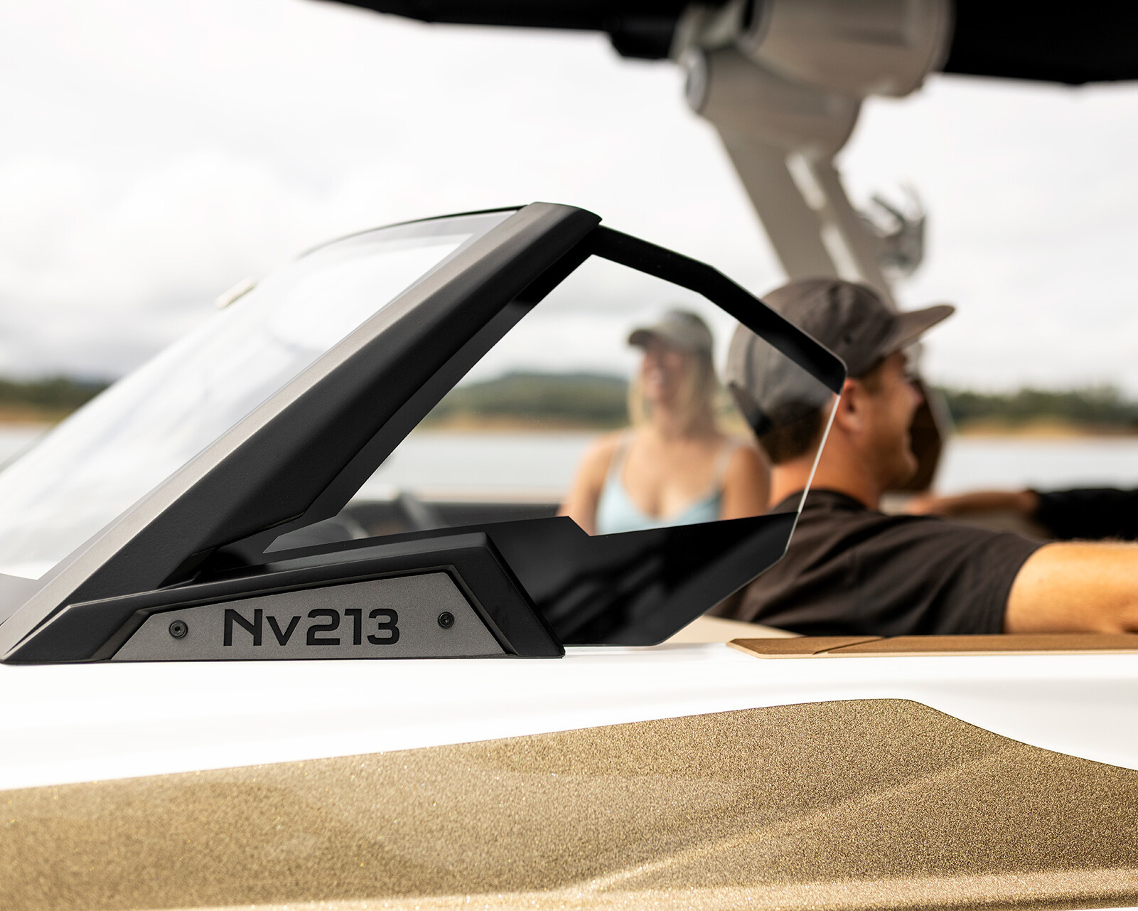 Close-up of a Centurion Nv213 windshield, with three people sitting inside—two in hats—enjoying a sunny day near the water.