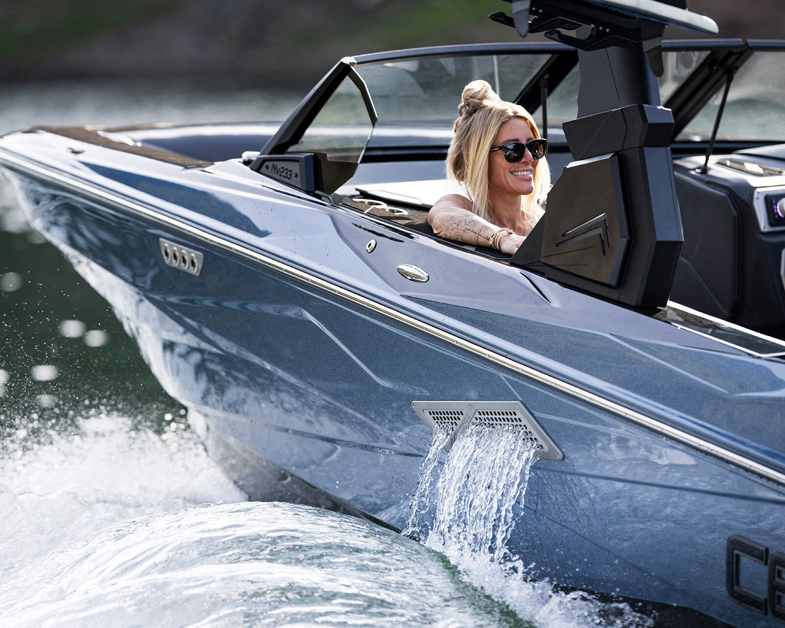 A woman wearing sunglasses drives a shiny blue Centurion Nv233 wake boat on the water, with spray and wake visible behind the boat.